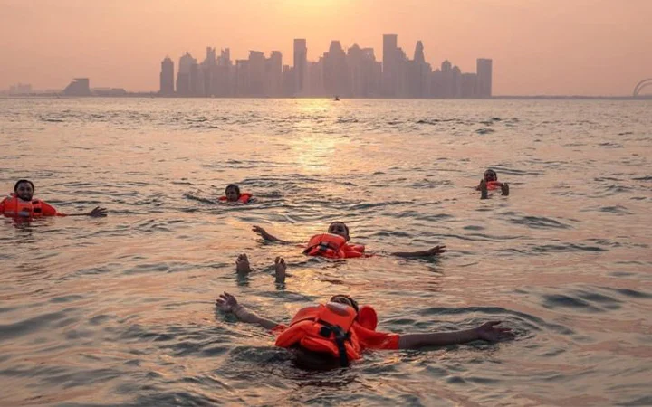 Five people wearing orange life jackets float on their backs in calm water at sunset during an Eid-Swim, with a city skyline visible in the background.