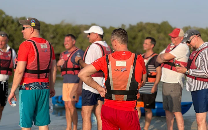 A group of men wearing life jackets stand together outdoors by water, some smiling and talking, with greenery and a kayaking sunrise in the blue sky background.