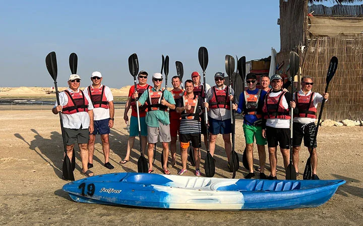 A group of twelve people in life jackets stand on a sandy shore at sunrise, holding kayak paddles upright behind a blue and white kayak, with rustic buildings and a clear sky in the background—a perfect start to a kayaking adventure.