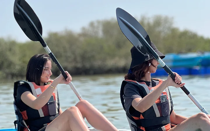 Two women wearing life jackets and holding paddles are kayaking on calm water at sunrise, with trees and blurred blue objects in the background under a clear sky.