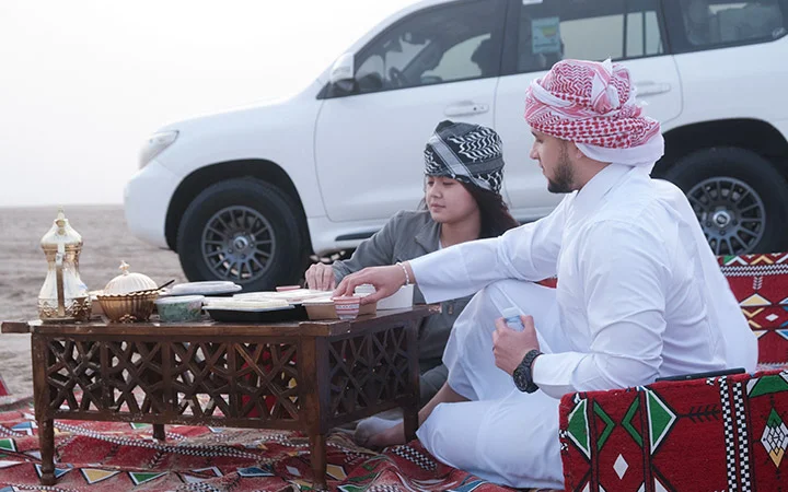 Two people sit on patterned cushions outdoors beside a white SUV, enjoying a Private Set-Up with traditional Arabic coffee and snacks on a low table in the desert. Both wear keffiyehs; one is dressed in white traditional clothing.