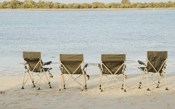 Four empty camping chairs form a private set-up on the sandy beach, facing calm waters and distant trees beneath a clear sky.