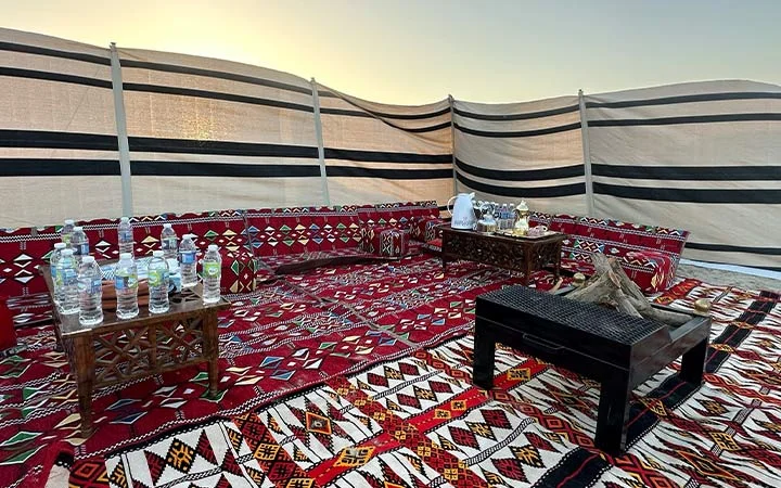 Traditional Middle Eastern outdoor private set-up with patterned red carpets, low wooden tables holding water bottles and drinks, and a tea set. Surrounded by striped fabric walls, under an open sky at sunset.
