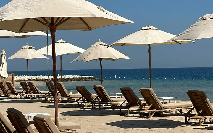 Rows of empty lounge chairs and large beige umbrellas line the sandy beach facing a calm blue sea, with a stone breakwater beyond—perfect for relaxing after an adventurous morning of quad biking and enjoying your included lunch access.