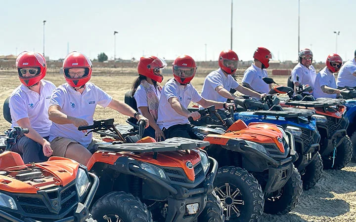 A group of people wearing red helmets and white shirts sit on orange and blue quad biking ATVs, lined up outdoors on a dry, open field under a clear sky, ready for adventure with included lunch access.