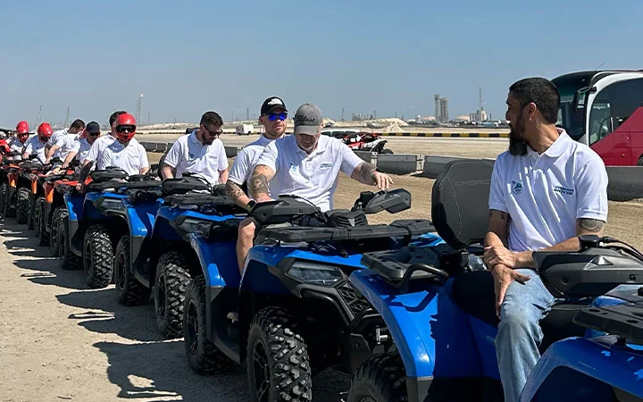 A group of people wearing white shirts and helmets sit on blue ATVs, ready for a thrilling quad biking adventure outdoors on a sunny day, with a bus, industrial buildings, and convenient lunch access in the background.