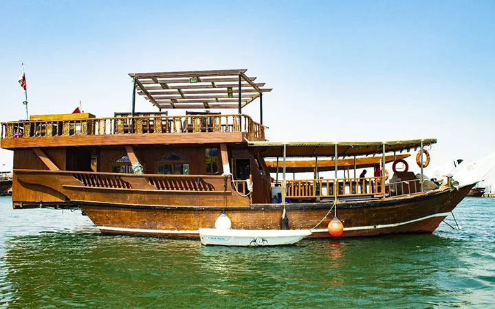 A traditional wooden dhow boat with two decks floats on calm water under a clear blue sky. Perfect for a Swim & Cruise - Dhow Cruise, it features orange lifebuoys and a small white dinghy tied to the side.