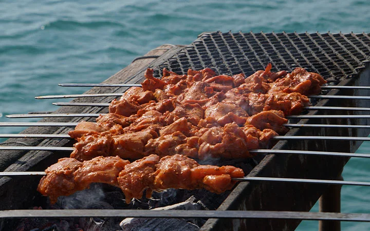 Several metal skewers with marinated meat are grilling over hot coals on a barbecue, while the ocean sets the perfect backdrop—ideal for a relaxing Swim & Cruise - Dhow Cruise experience.