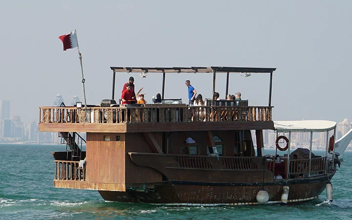 A traditional wooden boat sets sail on the sea for a Dhow Cruise. Several people enjoy the upper deck under a clear sky, while the Qatar flag waves at the stern and a city skyline provides a stunning backdrop.