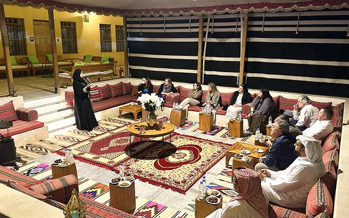 A group of people sit on red patterned cushions in a traditional tent, listening to a woman in black abaya speak during a Qatari Cultural Session. The setting features Arabic decor, rugs, and wooden tables with refreshments.