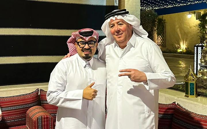 Two men wearing traditional Middle Eastern attire smile for the camera during a Qatari Cultural Session. One gives a thumbs up while the other flashes a peace sign, standing closely together among patterned cushions and lanterns outdoors.