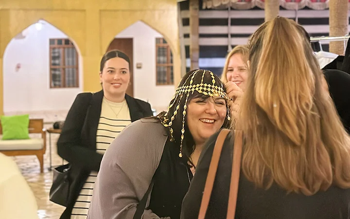 Four women indoors are smiling and laughing during a Qatari Cultural Session. One wears a decorative headdress with beads, while another adjusts it. The friendly atmosphere is enhanced by warm lighting and arched windows in the background.