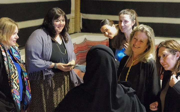 A group of women stand together in conversation, smiling and appearing engaged at a Qatari Cultural Session. One woman in the foreground wears a black hijab while others wear Western-style clothing. The setting features striped walls and patterned cushions.