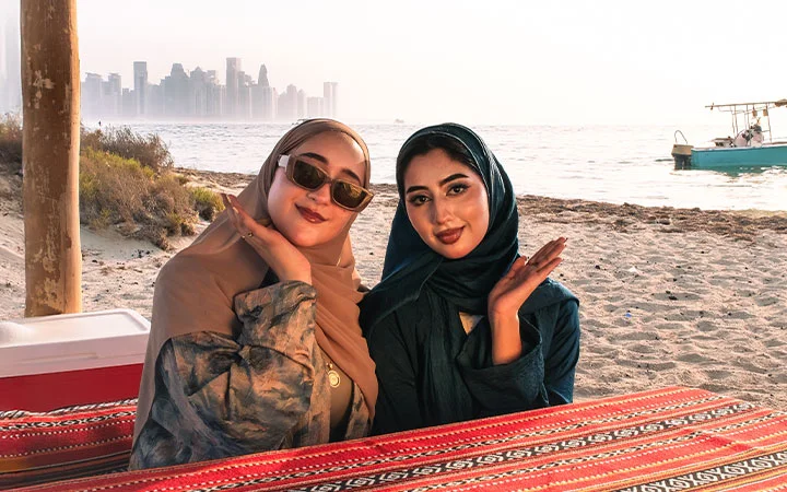 Two women in hijabs smile at a beachside table with a red patterned cloth, enjoying views of Safliyah Island from Doha. Behind them, the city skyline, a boat, and the sea stretch out beneath a hazy sky.