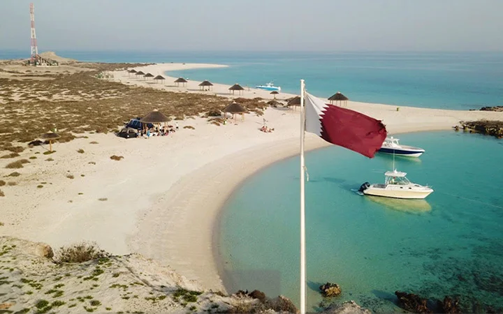 A Qatari flag waves near a curved sandy beach with clear turquoise water at Awa Island, where two anchored boats, people relaxing under umbrellas, and a distant antenna tower create the perfect luxury Day-Cation on a sunny day.