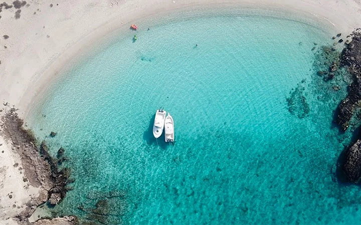 Aerial view of two boats anchored in clear turquoise water near Awa Island’s sandy beach, perfect for a luxury day-cation. Rocky shoreline and a few people lounging on the sand enhance this idyllic escape.