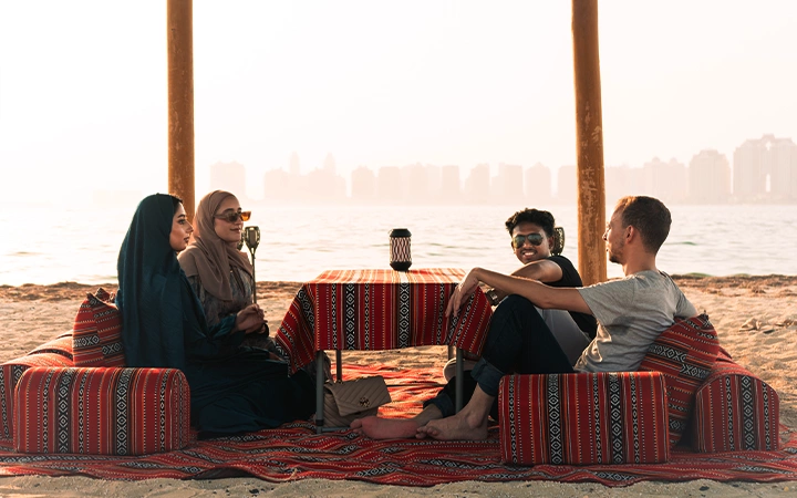 Four people sit on patterned cushions around a low table on a beach at sunset, with the Safliyah Island from Doha skyline and the sea in the background. Two women wear headscarves, and all appear relaxed and engaged in conversation.