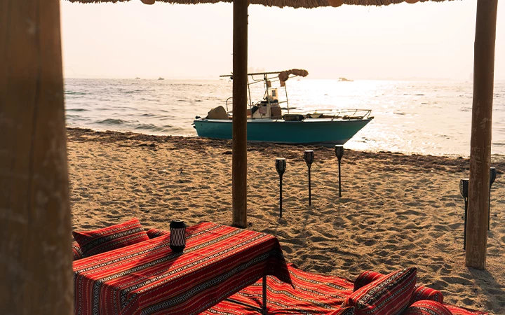 A small boat floats on calm water near a sandy beach at sunset. Patterned cushions and lanterns are arranged under a thatched shade, creating a relaxed seating area—perfect for enjoying the tranquil views of Safliyah Island from Doha.