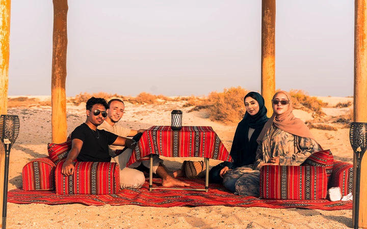 Four people sit on patterned cushions around a low table covered with a red cloth, under a canopy in a desert scene reminiscent of Safliyah Island from Doha. Two women wear headscarves; the group is smiling and relaxed, with sand and shrubs beyond.