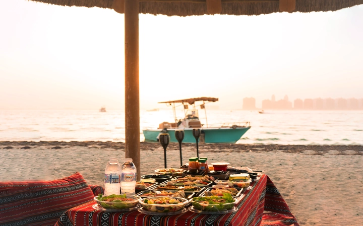 A table set with various dishes, two water bottles, and glasses sits under a thatched umbrella on a beach at sunset, overlooking Safliyah Island from Doha, with a boat drifting by and city buildings in the hazy background.