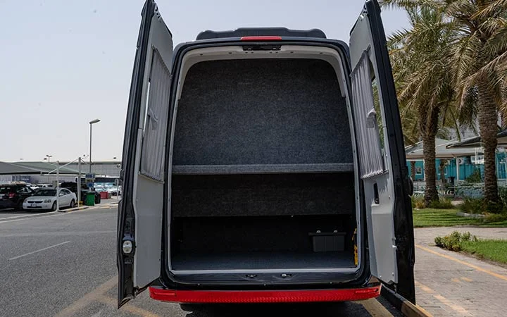 The image shows the rear view of an open white Mercedes Sprinter VIP Bus in Doha, with both back doors wide open, revealing an empty, carpeted cargo area with a shelf inside. Palm trees and cars are visible in the background.