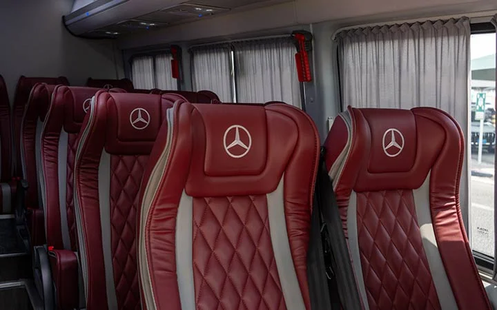 Rows of maroon, quilted-leather seats with Mercedes-Benz logos on headrests inside a modern Mercedes Sprinter VIP Bus Doha; gray accents and closed curtains on the windows are visible in the background.