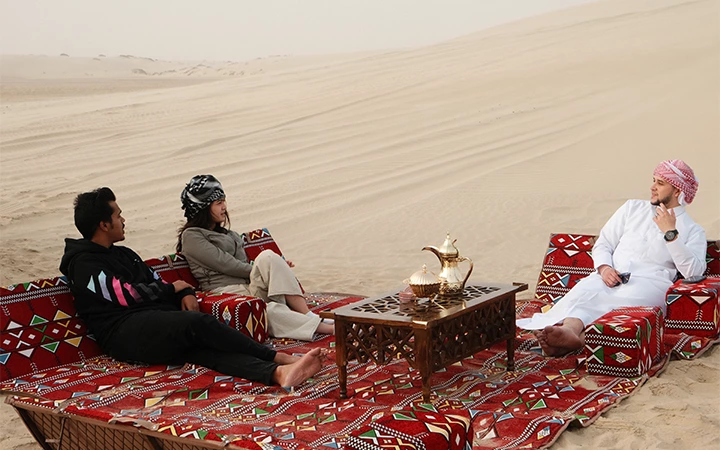 Three people sit on patterned cushions around a low table with a teapot and cups, relaxing in a sandy desert under Qatar’s breathtaking sunrise. Two wear casual clothes, while one is dressed in a traditional white robe and headscarf.