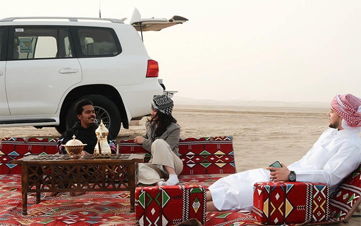 Three men sit on colorful patterned cushions and rugs in the desert beside a white SUV, sharing a traditional tea set on a low wooden table. Qatar’s breathtaking sunrise peeks above the horizon, illuminating sand stretching into the distance.