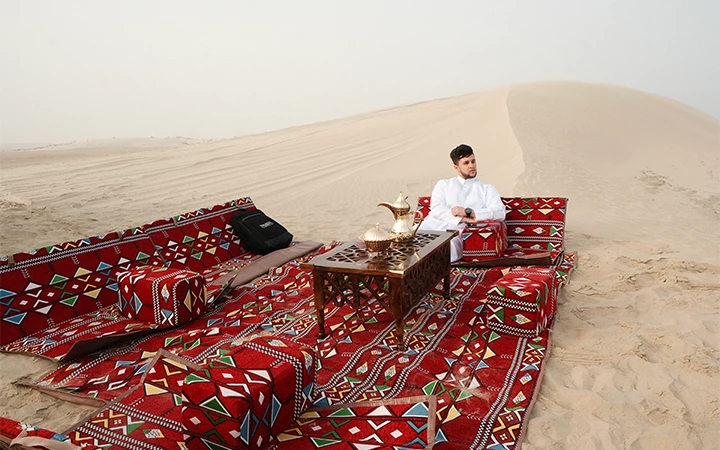 A man in traditional white clothing sits on patterned red cushions around a low table with tea sets, arranged on sand as Qatar’s breathtaking sunrise illuminates the desert and a large dune rises in the background.