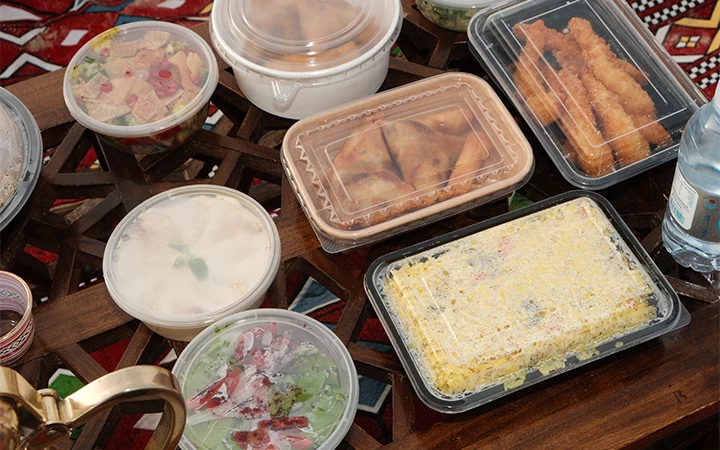 A variety of takeout food containers with assorted dishes—including salad, rice, fried snacks, and soup—are arranged on a wooden table with a patterned tablecloth underneath, reminiscent of enjoying breakfast under Qatar’s breathtaking sunrise.