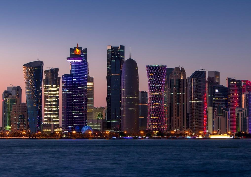 Nighttime cityscape of Doha, Qatar, featuring modern skyscrapers with colorful lights reflecting off the buildings, viewed from across the water under a darkening sky.