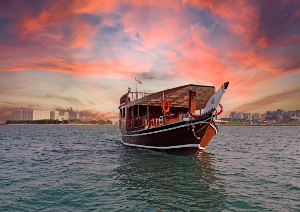 A traditional wooden boat floats on calm water at sunset, with a dramatic pink and orange sky and a city skyline in the distant background.