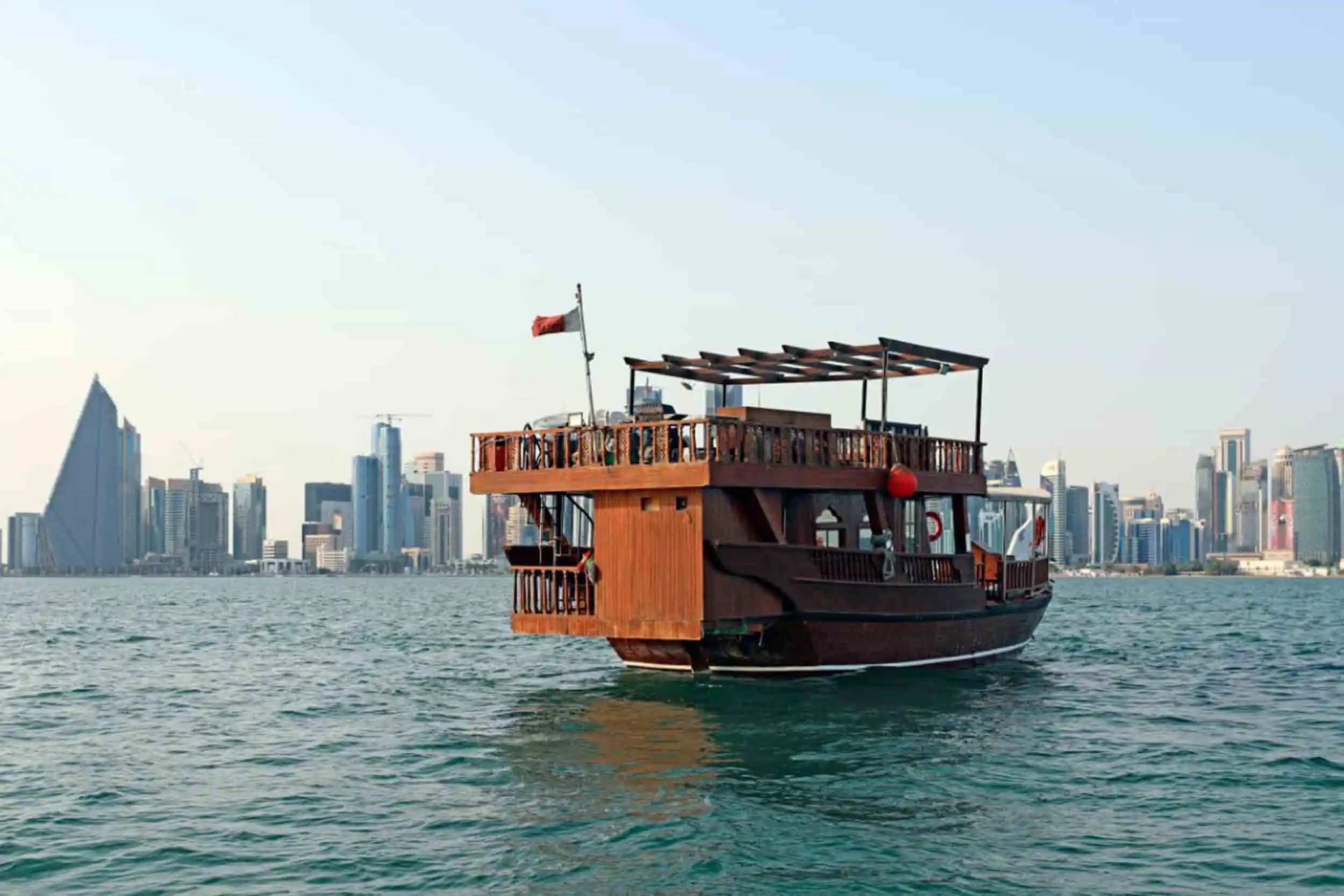 A traditional wooden dhow boat floats on the water with a city skyline and modern skyscrapers in the background under a clear sky.