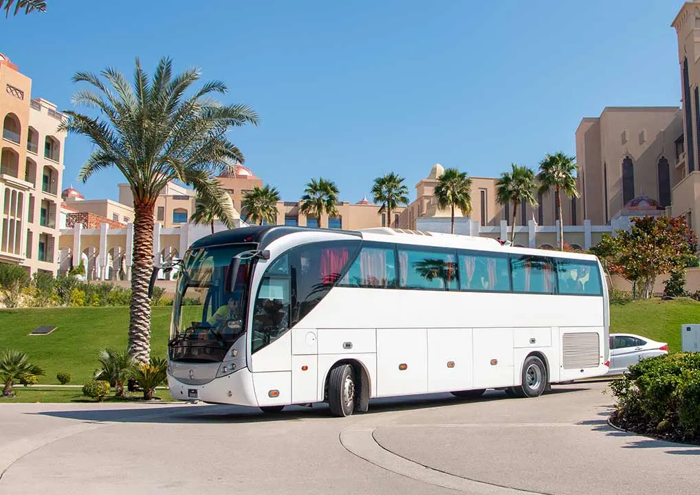A white tour bus is parked on a circular driveway surrounded by palm trees and modern beige buildings under a clear blue sky.