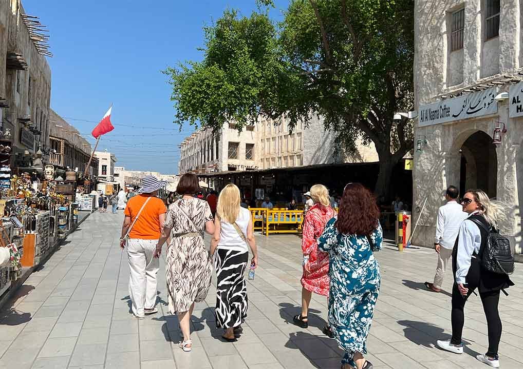 A group of people walk along a sunny, open-air market street lined with shops and stalls, with a tree and a building in the background and a red flag on the left.
