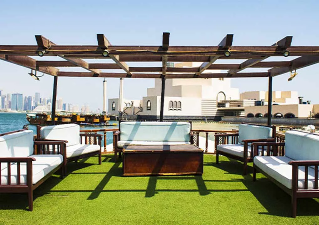 Outdoor patio with wooden pergola, cushioned benches, and a wooden table on green artificial turf, overlooking water and modern city buildings under a clear sky.