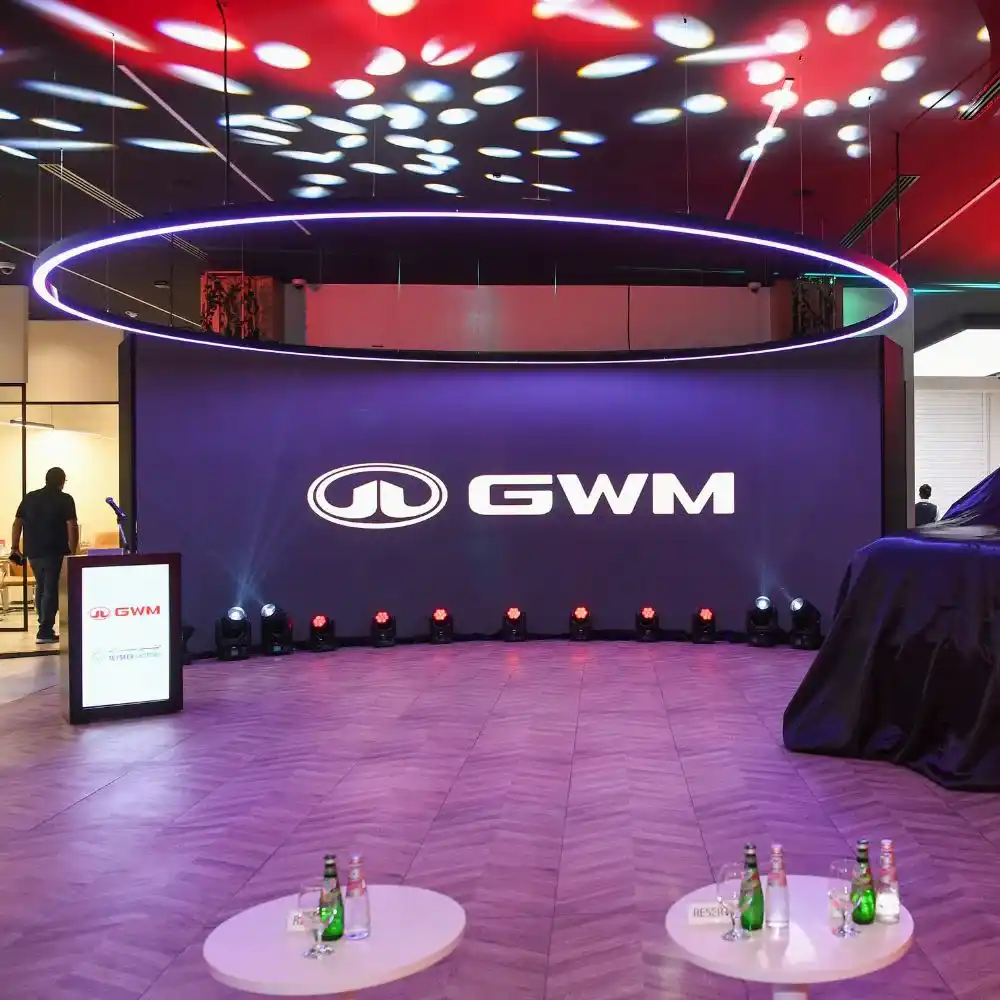 A modern showroom features a large LED screen displaying the GWM logo. Spotlights and circular ceiling lights add ambience. In the foreground, tables with water bottles are visible. People are present in the background.