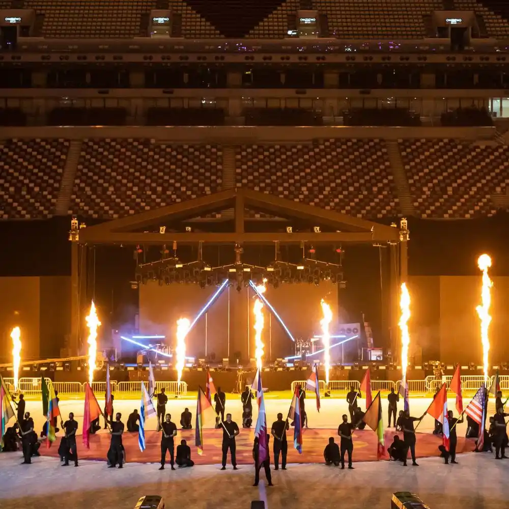 Performers hold various national flags on a stadium field with rows of empty seats behind them. Flames shoot up from the stage, and bright lights create dramatic effects during the event.