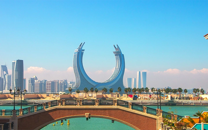 A view of the Katara Towers, two curved skyscrapers in Lusail, Qatar, set against a clear blue sky. Water, palm trees, and a stone bridge enhance the scene—a must-see on any Shared Doha City Tour, with modern buildings in the background.