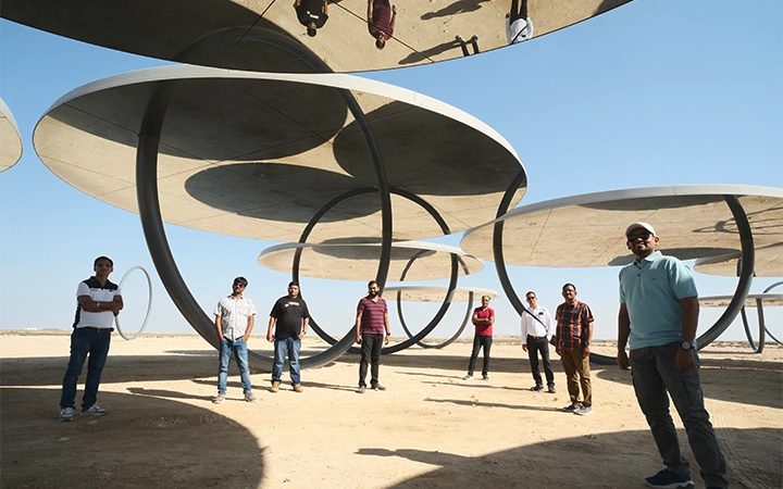 A group of people on a Shared Doha City Tour stand under large, circular, modern art structures that cast shadows on sandy ground, with clear blue sky above. Some reflections of people are visible on the shiny undersides of the sculptures.