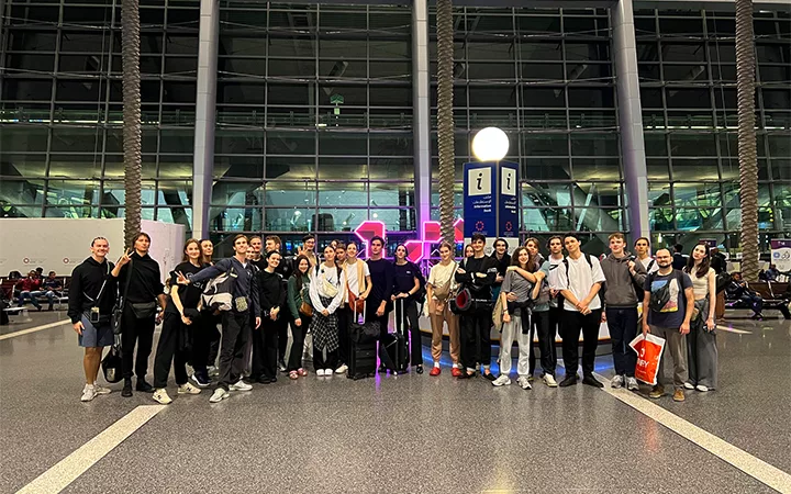 A large group of people stands together posing for a photo inside a modern airport terminal, likely before embarking on their Shared Doha City Tour, with tall glass windows, palm trees, and illuminated signs in the background.