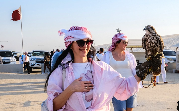 A smiling woman in sunglasses and a traditional headscarf holds a falcon on her gloved hand during a Shared Doha City Tour, with other people, cars, and a red-and-white flag visible in the sunny desert backdrop.