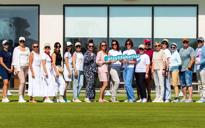 A group of people stand in a row on a lawn, smiling at the camera during a Shared Doha City Tour. Two people in the center hold a blue sign that says playlifebetter, with a modern building featuring large windows in the background.