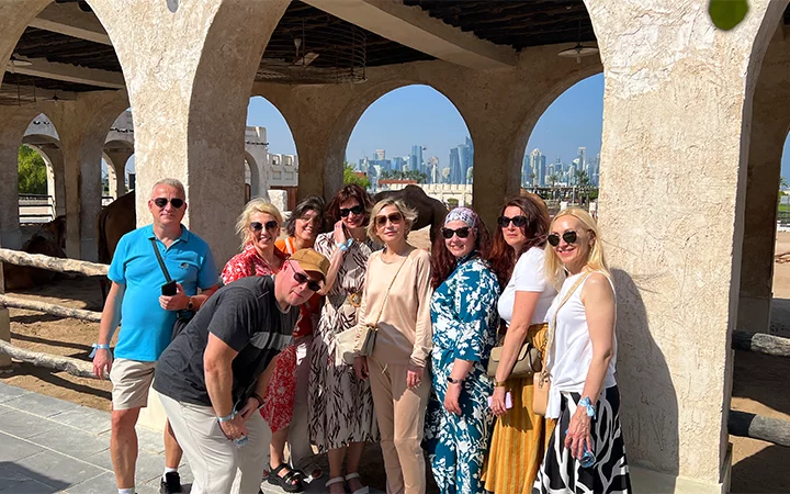 A group of nine adults poses and smiles under a stone archway with a city skyline and blue sky in the background. Enjoying a sunny day in summer clothes, they capture the fun of their Shared Doha City Tour.