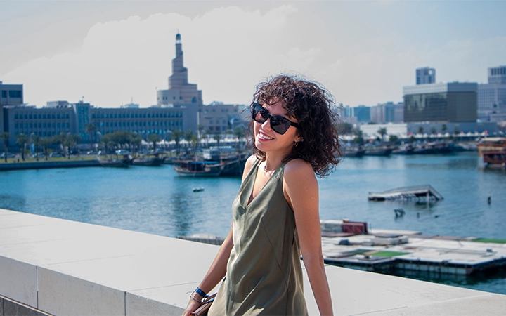 A woman wearing sunglasses and a sleeveless dress smiles while standing by a waterfront, perhaps enjoying a Shared Doha City Tour. City buildings and boats are visible in the background under a clear sky.